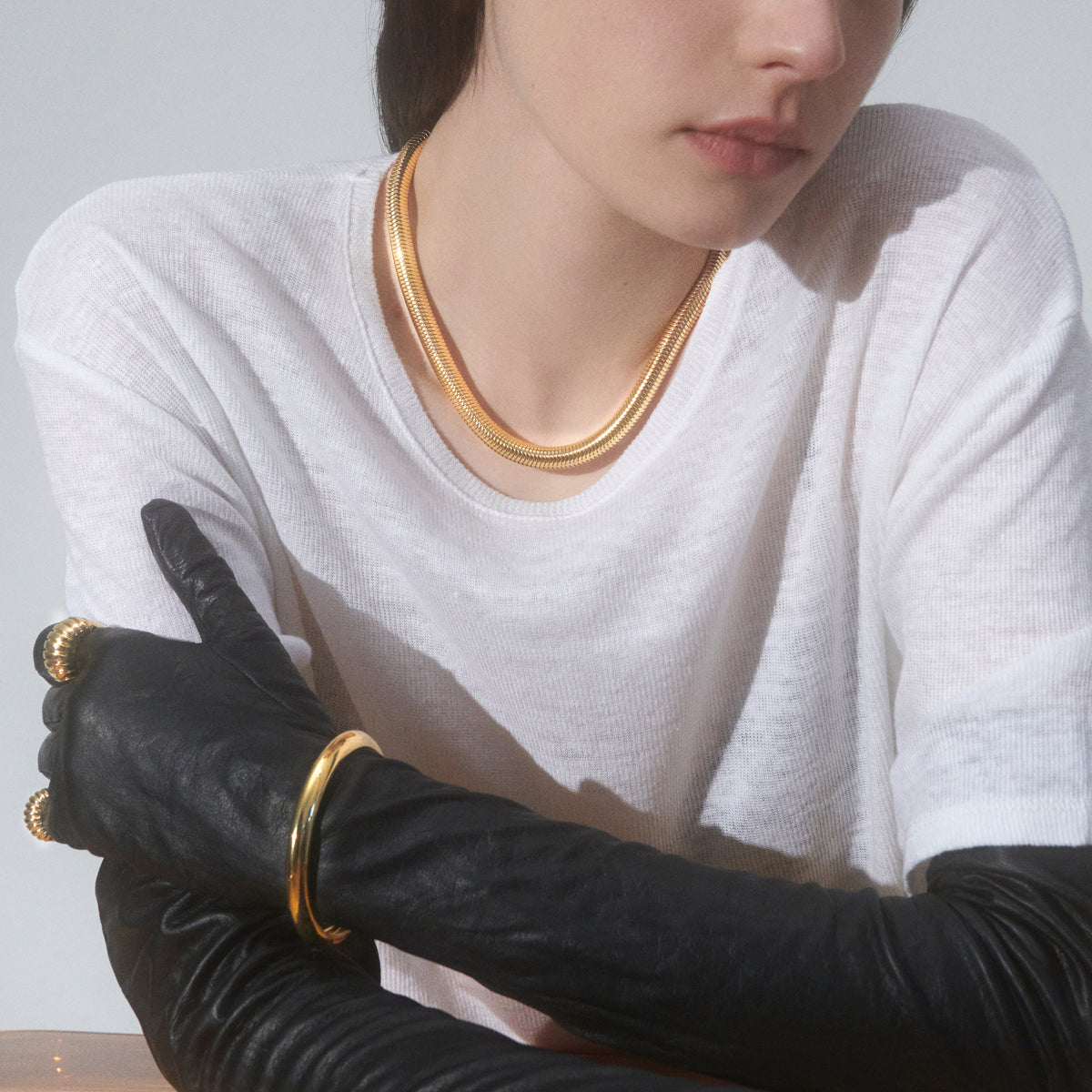 A close-up of a model wearing a layered gold chain necklace, a gold bangle bracelet, and black leather opera gloves over a white long-sleeve shirt.