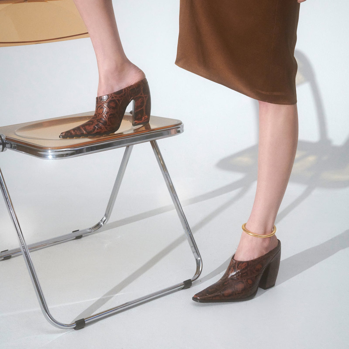 A person wearing brown snakeskin-patterned pointed-toe mule heels with block heels, seated on a modern metal chair against a white studio background.